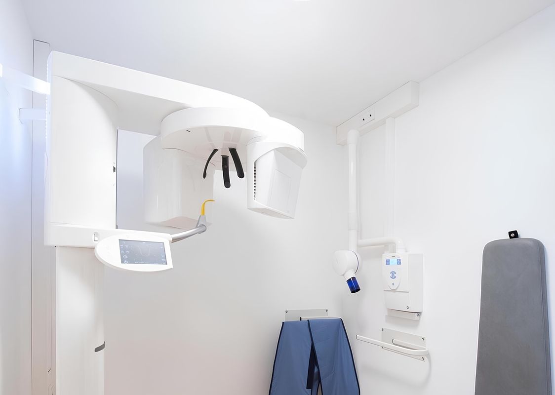 Moderne zahnmedizinische Praxis mit einem weißen Röntgengerät, einem Monitor und medizinischen Instrumenten, sowie einem blauen Schutzkleid und einem Stuhl im Hintergrund